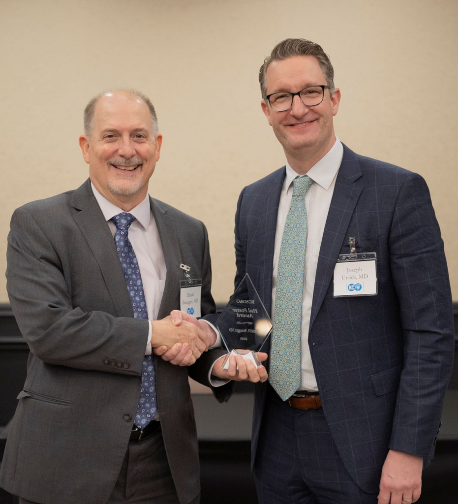 Two men in suits shake hands, one holding an award in a room with a light wall.