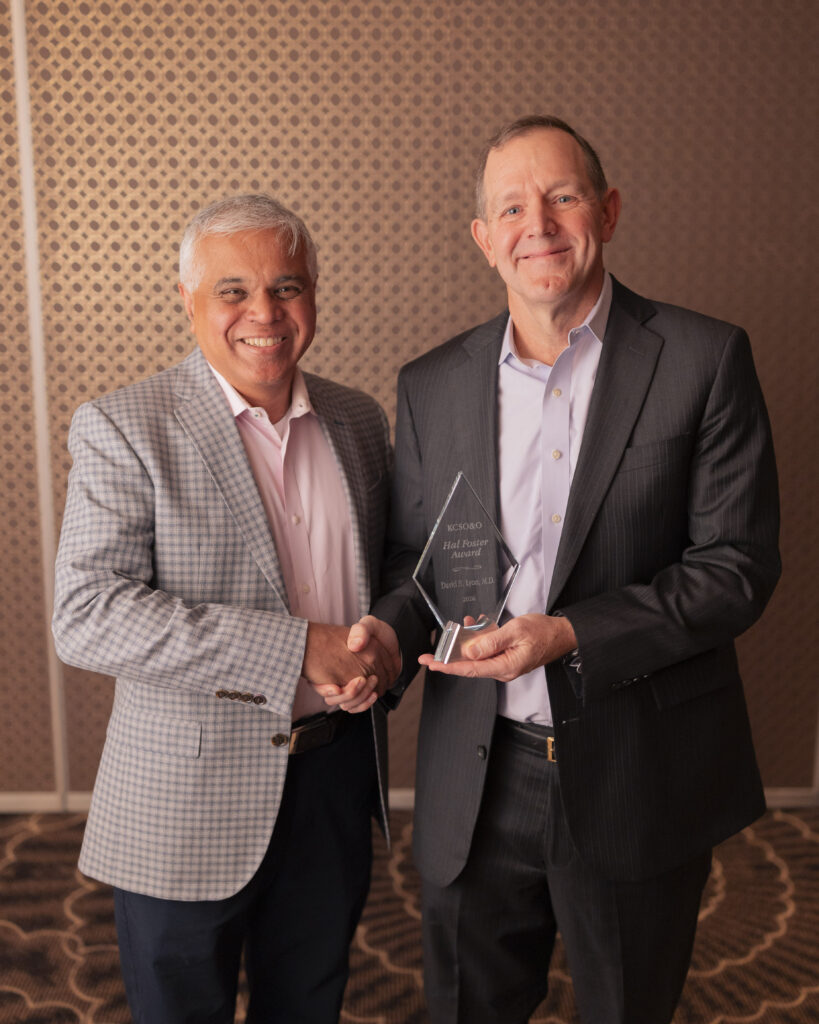 Two men in suits shake hands, one holding an award in a room with a patterned wall.