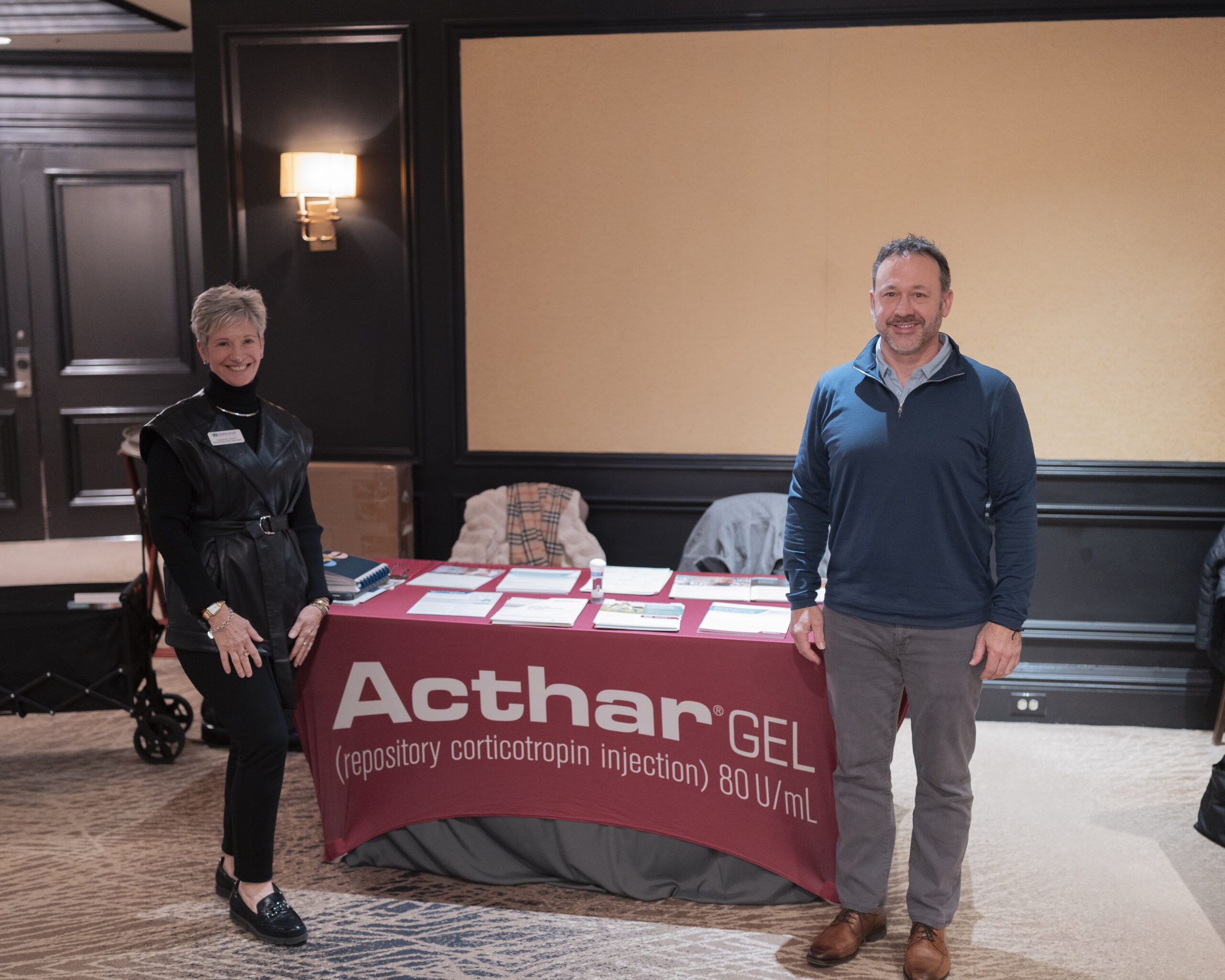 A woman and a man standing to the side and in front of the exhibitor table for Acthar Gel